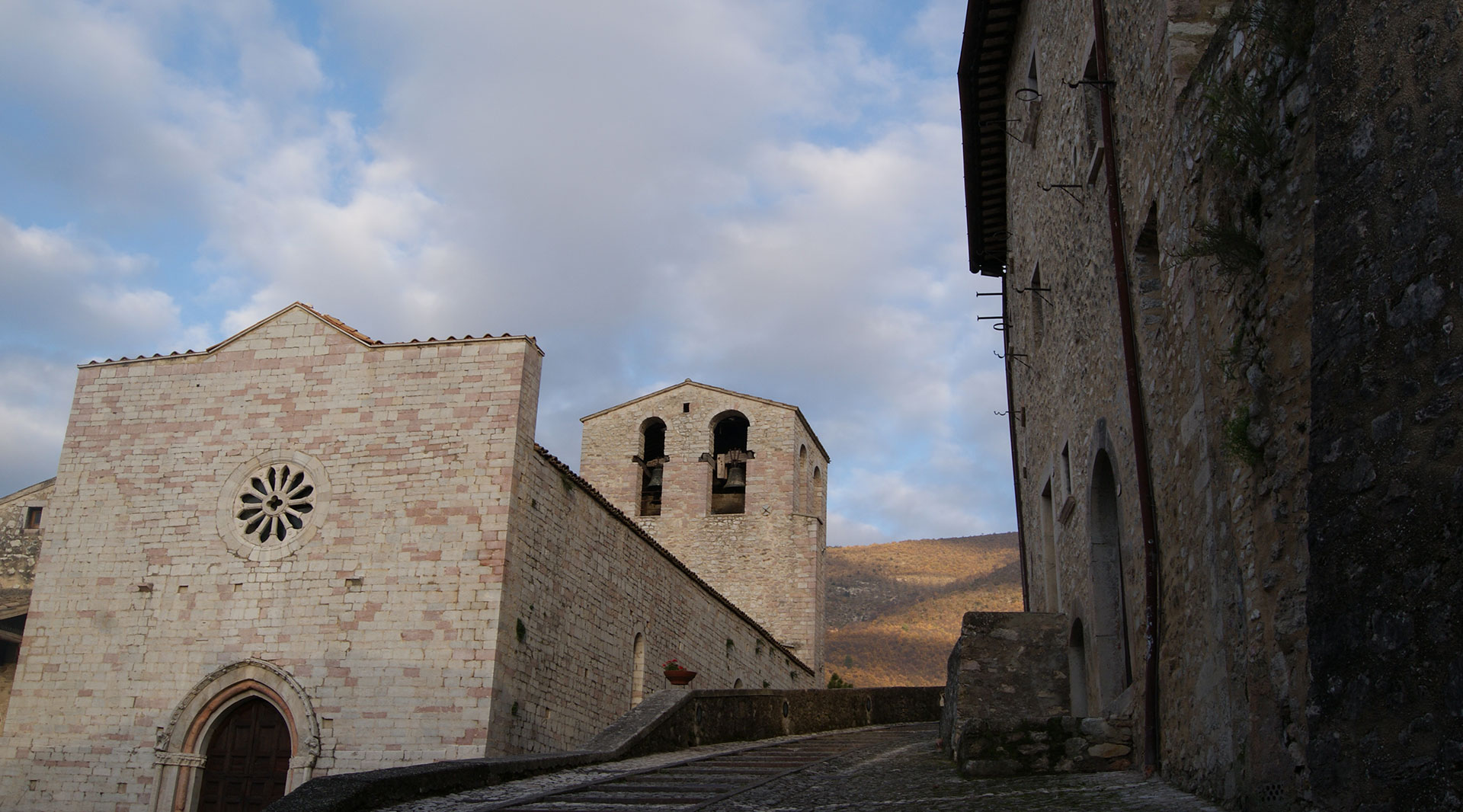 Church of Santa Maria Assunta in Vallo di Nera, with a stone façade, rose window, and bell tower, nestled in a hilly landscape.