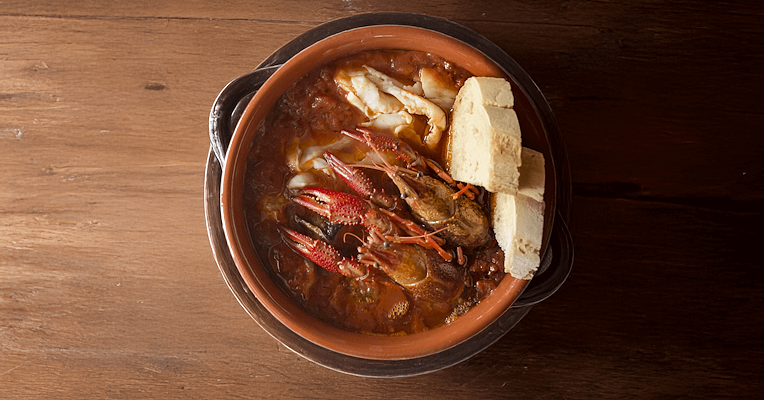 Tegamaccio di pesce di lago con gamberi di fiume, servito in una pentola di terracotta con fette di pane, su tavolo di legno.