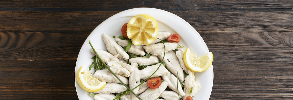 Perch fillets served with rocket, cherry tomatoes, chives, and lemon slices on a white plate, placed on a wooden table.