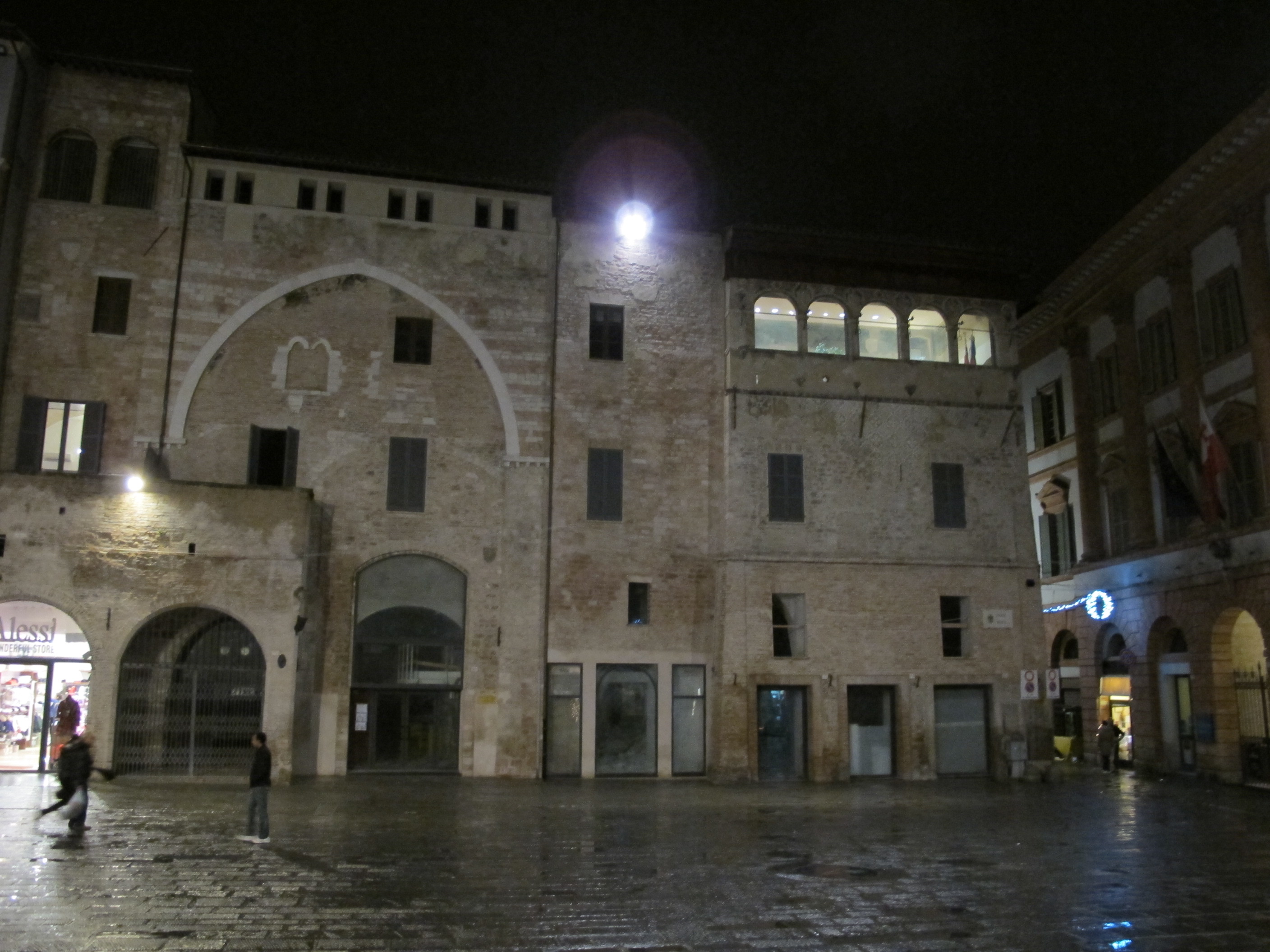 Edificio storico in mattoni con arco ogivale e loggia illuminata, di notte, in una piazza.