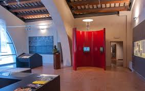 Sala museale con teche espositive, pannelli informativi, una struttura rossa centrale e soffitto con travi in legno a vista.