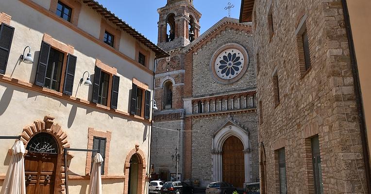 Blick auf einen Platz mit Gebäuden aus Stein und Ziegel, dunklen Fensterläden und einer Kirche mit Rosette und Glockenturm im Hintergrund.