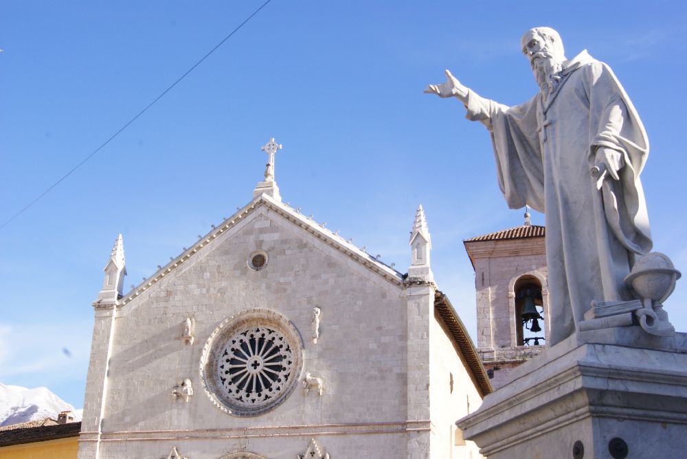 Statue of Saint Benedict in Norcia, with the Basilica in the background, its Gothic rose window, and the bell tower under a clear sky.
