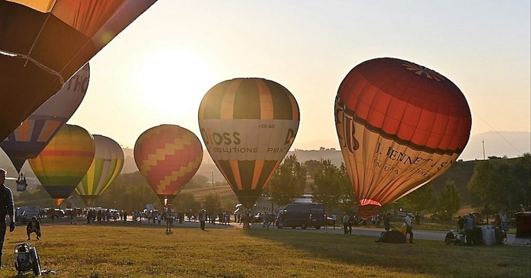 Eine Gruppe von Menschen bereitet den Start von Heißluftballons vor, die auf einer Wiese liegen, während die Sonne am Himmel aufgeht.