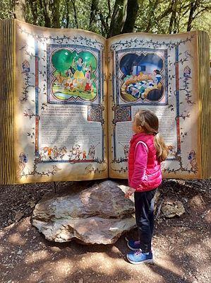A little girl is carefully reading a fairy tale written on a giant book placed on a boulder in a forest.