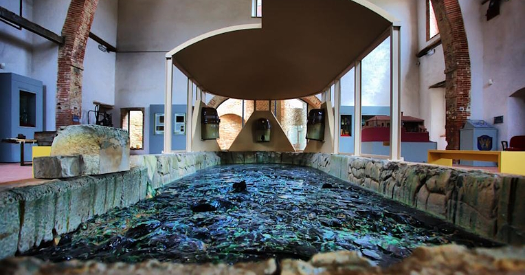 Interior of the Glass Museum in Piegaro, with a stone basin containing fragments of colored glass and exposed brick arches.