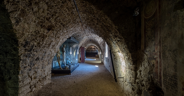 Un corridoio sotterraneo con pareti di pietra, teche contenenti mummie e antichi affreschi sbiaditi.