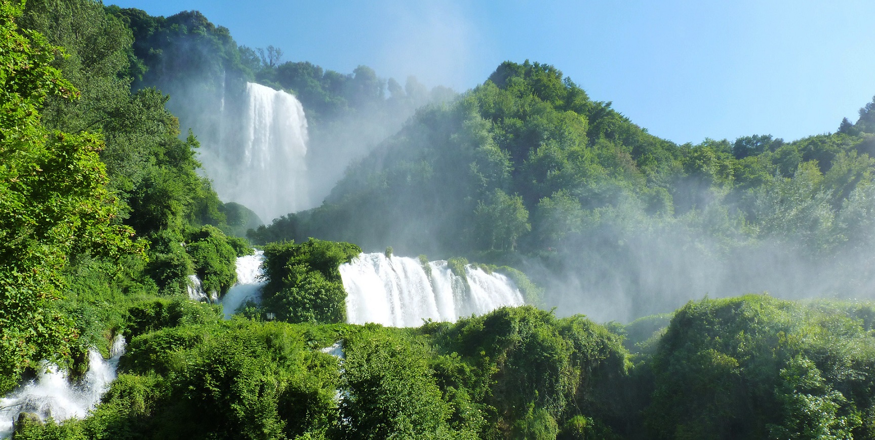 La Cascata delle Marmore vista dal basso: salti d’acqua tra fitte vegetazioni, spruzzi e vapore che si alzano verso il cielo limpido.