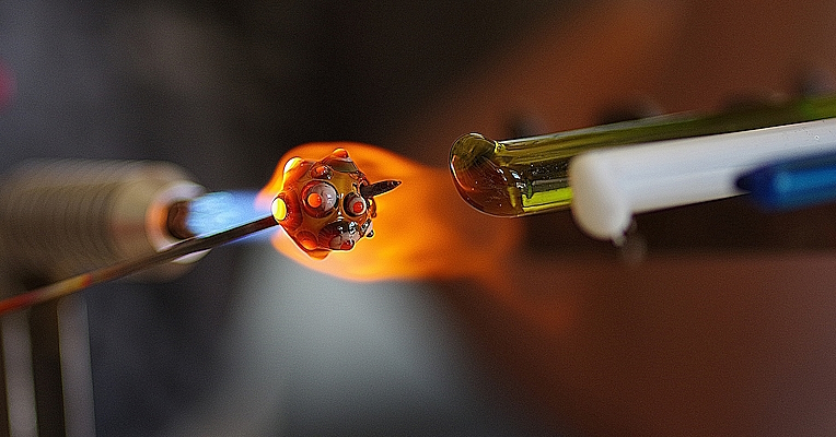 Bild mit Glasstäben in verschiedenen Farben und einer Glaskugel, die auf einen durch Feuer erhitzten Metallstab aufgefädelt ist