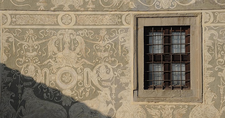 Wall decorated with white Renaissance motifs on a gray background and a barred window in the foreground.
