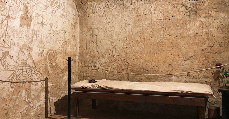 Walls engraved with symbols, crosses, and centuries-old inscriptions in a prison cell lit by a small barred window.