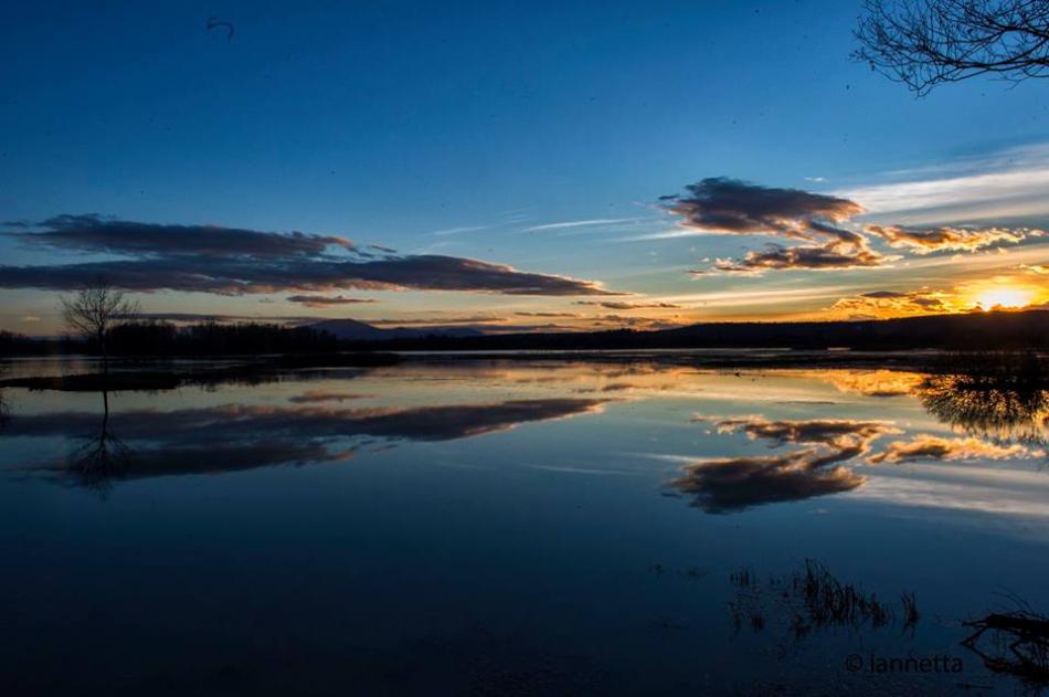 Sonnenuntergang über dem See mit goldenem Himmel und Wolken, die sich im ruhigen Wasser spiegeln, zwischen Bäumen und dunklen Hügeln.