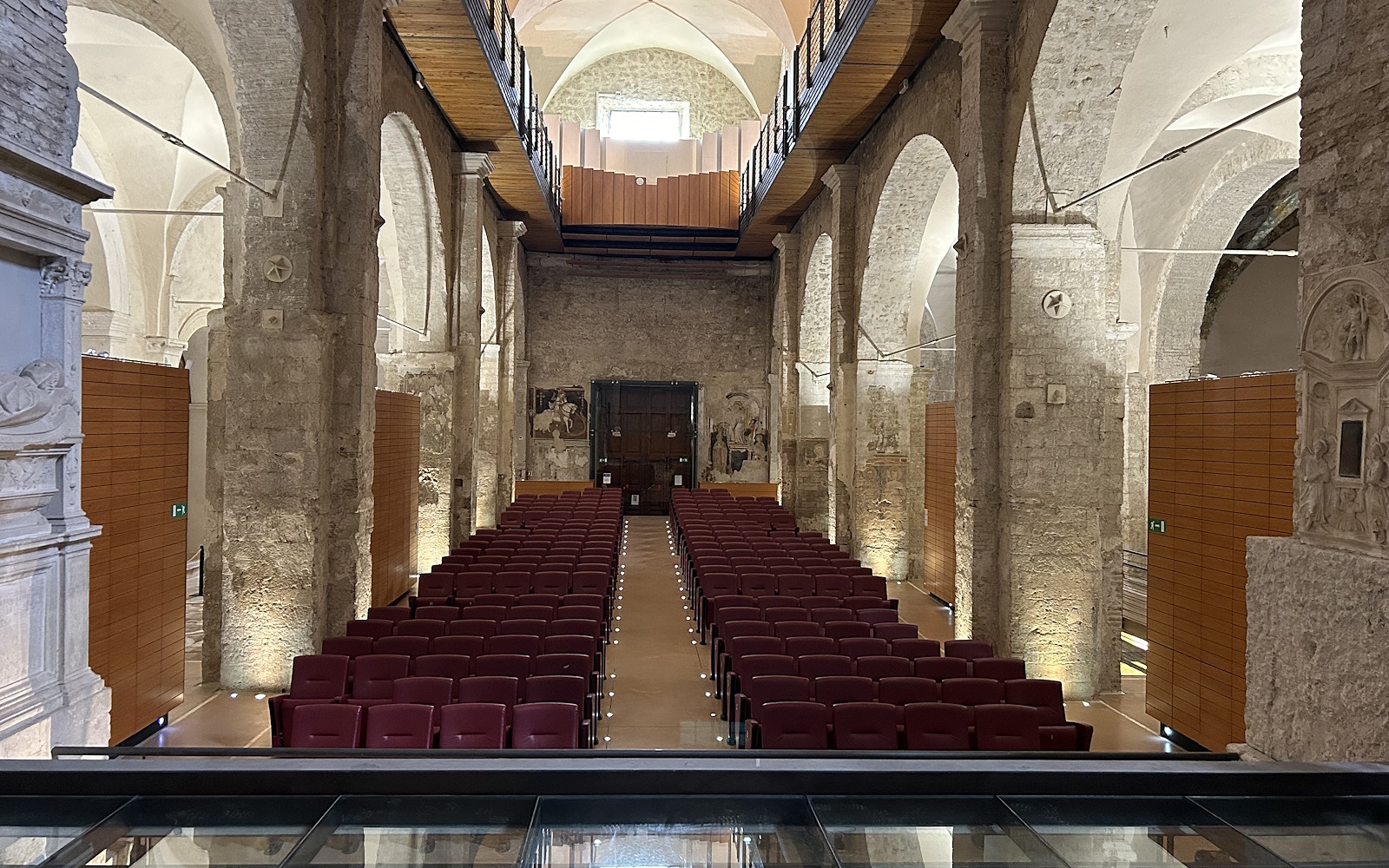 Interno dell’Auditorium Bortolotti a Narni con file di sedili rossi disposti simmetricamente, pareti in pietra, un soffitto con volte a crociera e una passerella in legno al piano superiore che corre lungo entrambi i lati della sala. Sullo sfondo, una grande porta di legno e affreschi parzialmente visibili adornano la parete.
