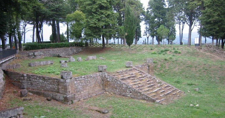Ruinen eines antiken Steingebäudes mit zentraler Treppe, umgeben von Bäumen und grüner Wiese.