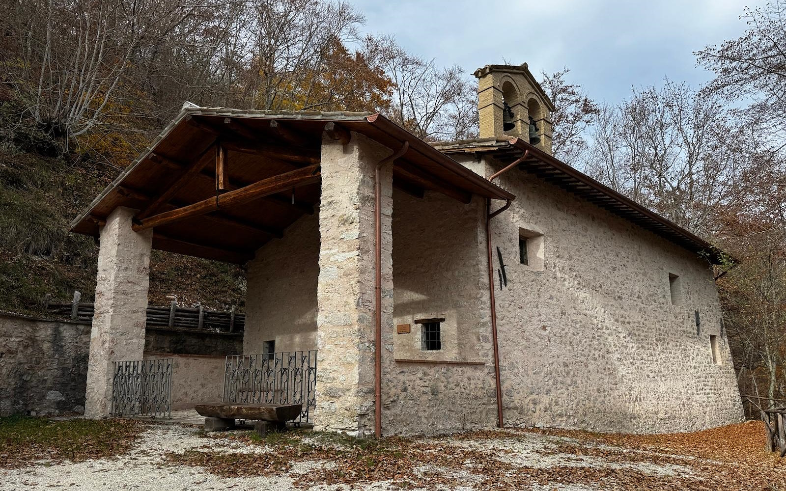 Piccolo eremo in pietra con campanile e portico in legno, immerso tra gli alberi su un pendio di montagna.