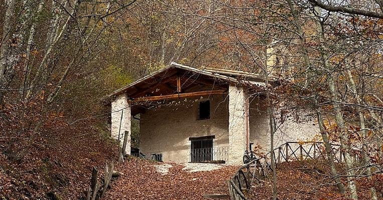 Piccolo eremo in pietra con campanile e portico in legno, immerso tra gli alberi su un pendio di montagna.