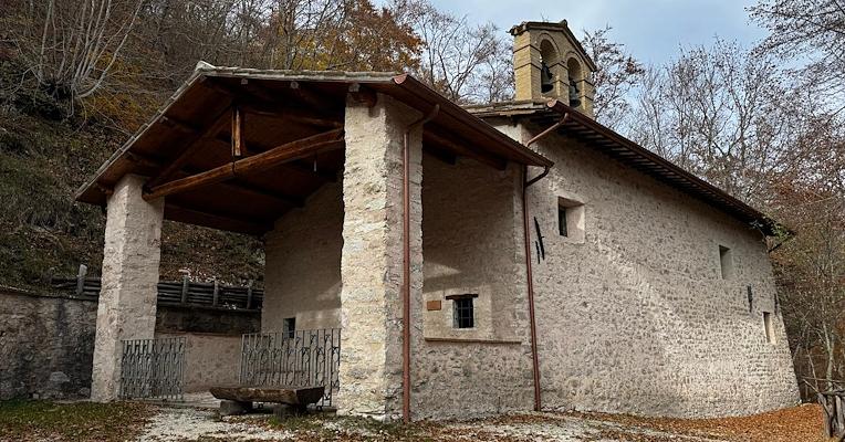 Piccolo eremo in pietra con campanile e portico in legno, immerso tra gli alberi su un pendio di montagna.