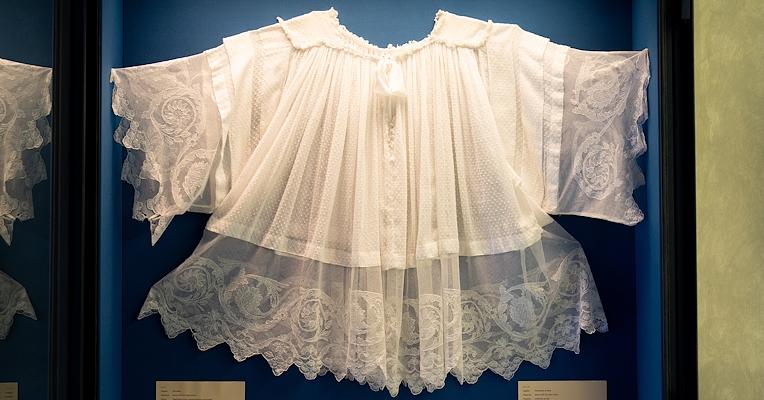 Abito da battesimo bianco ricamato in tulle, con maniche ampie e dettagli decorativi, esposto in una vetrina del museo.