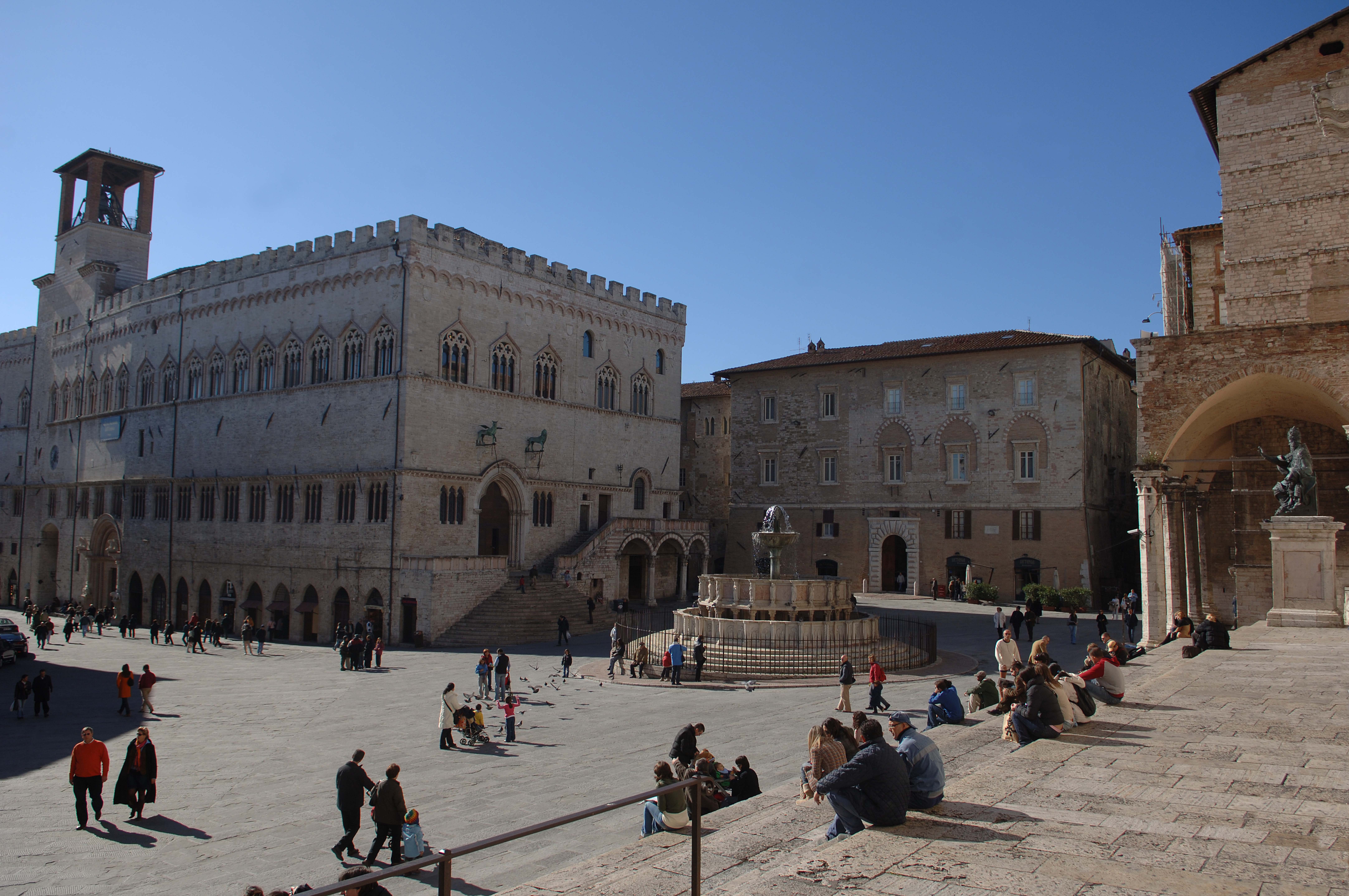 Piazza IV Novembre mit dem großen Brunnen und dem Palazzo dei Priori, Passanten genießen den sonnigen Tag auf den Stufen der Kathedrale sitzend oder schlendern entlang.