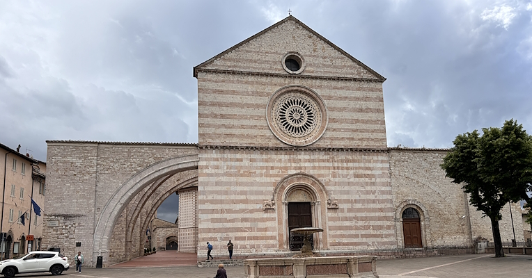 Facade of the Basilica of Santa Chiara in Assisi, featuring a rose window and side arch, viewed from the square with a fountain in the foreground.