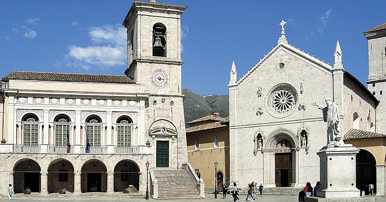 Central square with the town hall and the Basilica of San Benedetto in Norcia, featuring a bell tower and a statue in the foreground.
