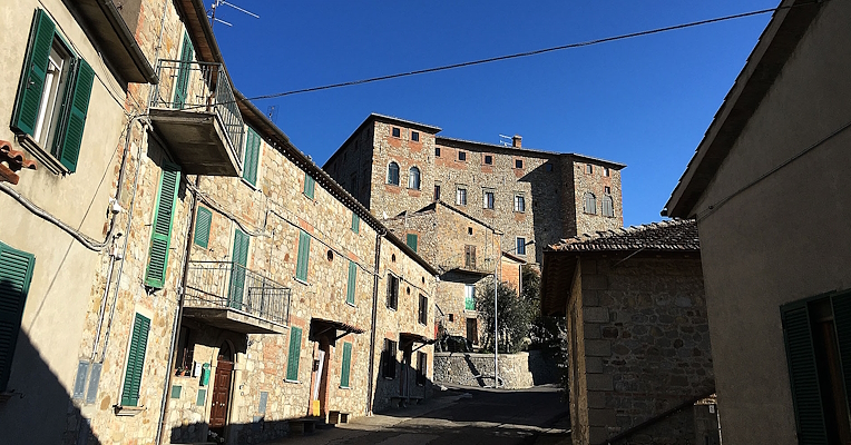 Street in the village of Carnaiola with stone houses and green shutters under a clear sky; a castle at the top of the road.