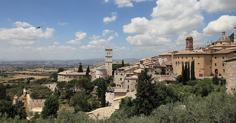 Panorama von Assisi mit historischen Steingebäuden, Glockentürmen und Türmen bei klarem Himmel, aber mit einigen Wolken