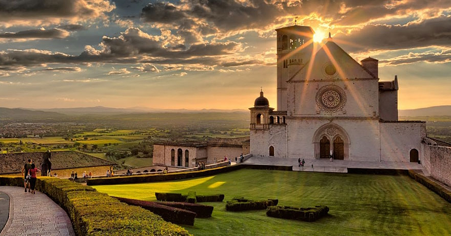 Die Basilika des Heiligen Francesco in Assisi bei Sonnenuntergang, wenn die Sonne durch die Wolken bricht und die Wiese und das Tal beleuchtet