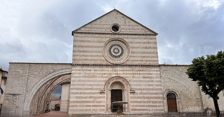 Fassade der Basilika der Heiligen Chiara in Assisi, mit ihrem charakteristischen rosa Stein und dem Brunnen im Vordergrund.