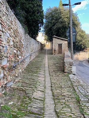 A stone and brick staircase with a side wall, bordered by a stone wall and trees, leads to a convent in the background.