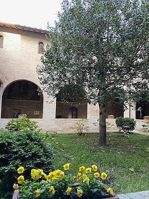 A cloister with a brick portico, a well-kept garden with yellow flowers, and a tree in the centre, in a serene and historical atmosphere.