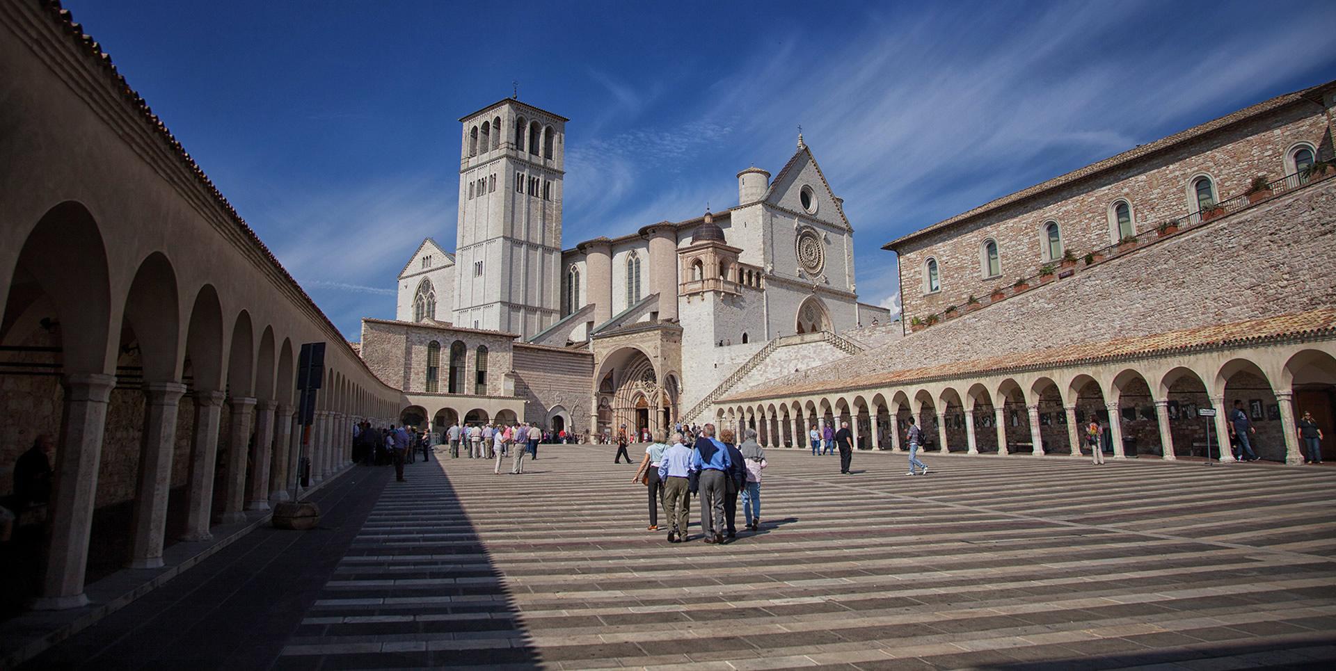 Piazza Inferiore di San Francesco ad Assisi, caratterizzata da portici laterali in pietra e dalla presenza della Basilica Superiore sullo sfondo. La scalinata centrale conduce all'ingresso della Basilica Superiore, evidenziando l'architettura gotica del luogo.