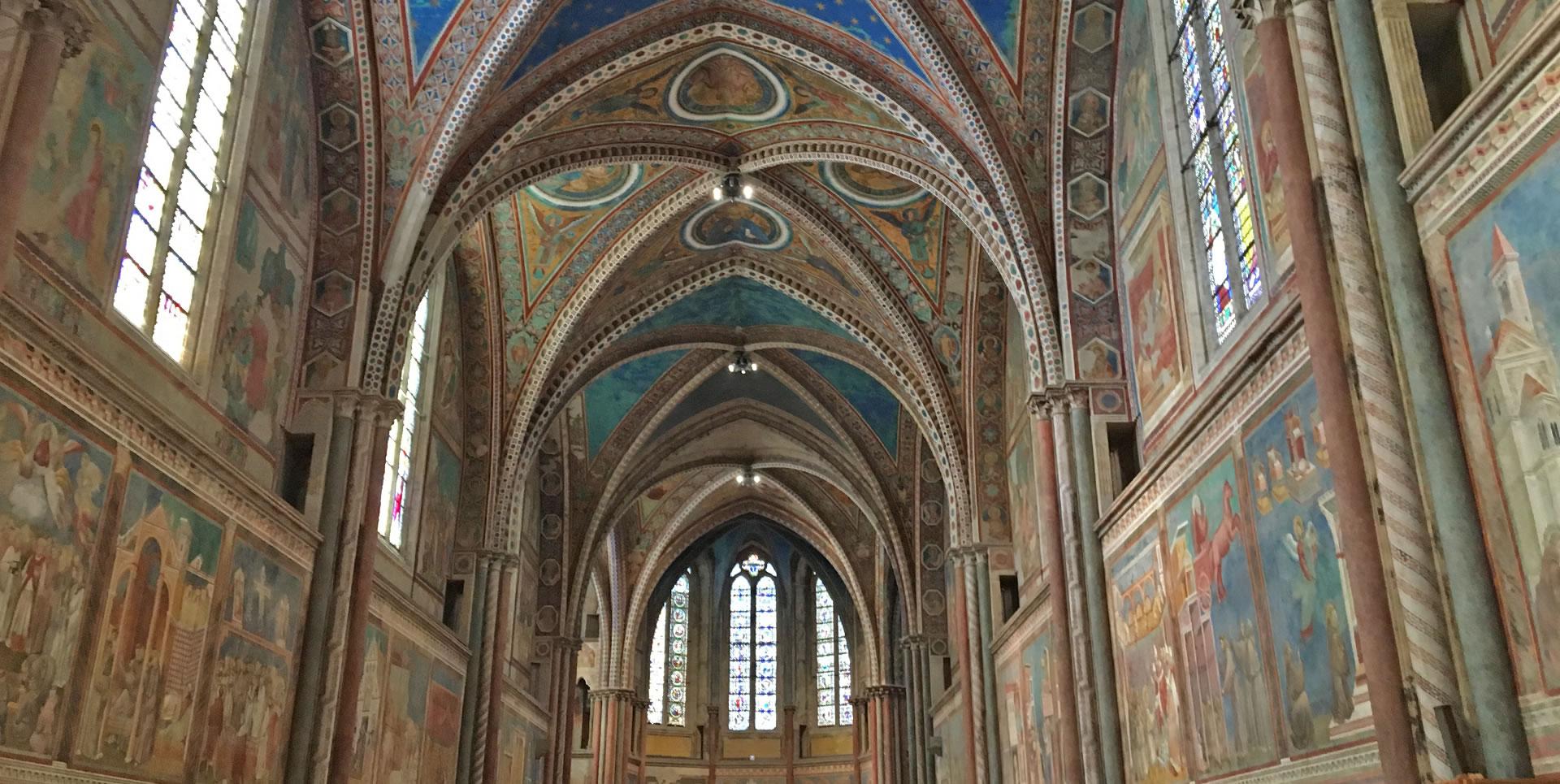 Vista interna della Basilica Superiore di San Francesco ad Assisi, con le sue alte volte a crociera decorate. Le pareti, ricoperte di affreschi realizzati da artisti come Giotto e Cimabue, rappresentano scene della vita di San Francesco e storie bibliche. Grandi vetrate permettono alla luce naturale di illuminare l'interno.