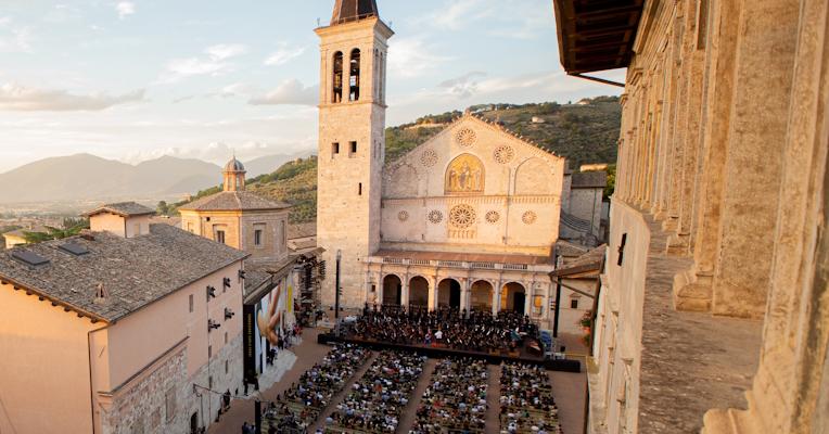 Vista dall’alto di Piazza Duomo a Spoleto durante una serata del Festival dei Due Mondi con il pubblico seduto di fronte al palco allestito davanti alla cattedrale romanica, illuminata dalla luce calda del tramonto, con sullo sfondo il paesaggio collinare umbro e la città sottostante.