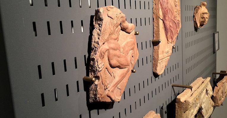 Frammenti di terracotta decorata con figure umane, esposti su una parete forata nera in un museo archeologico