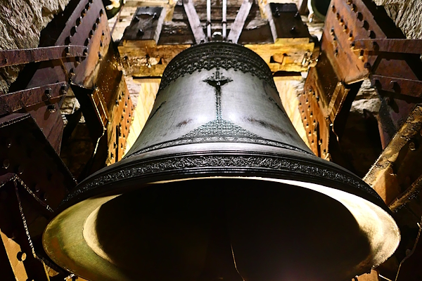 Ornate bronze bell seen from below, mounted in a wooden structure inside a bell tower