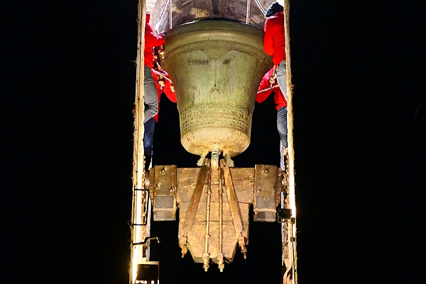 Ornate bronze bell seen from below, mounted in a wooden structure inside a bell tower