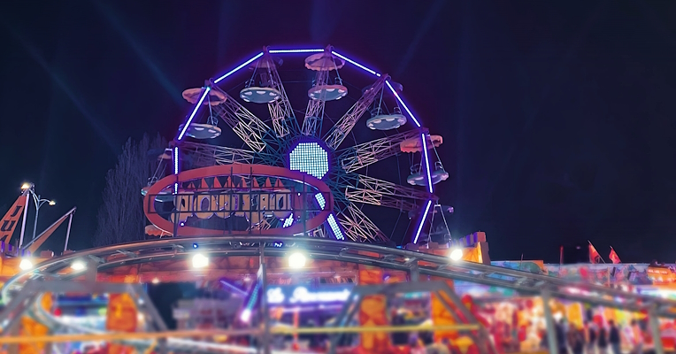 Night-time funfair with Ferris wheel lit by purple and blue lights, with colorful rides and stalls in the foreground