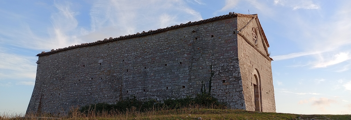 Veduta laterale di un edificio in pietra situato su un’altura erbosa, con cielo azzurro e nuvole leggere sullo sfondo.