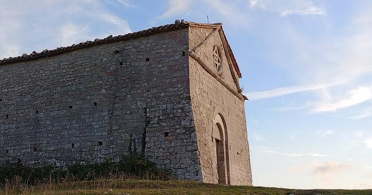 Veduta laterale di un edificio in pietra situato su un’altura erbosa, con cielo azzurro e nuvole leggere sullo sfondo.
