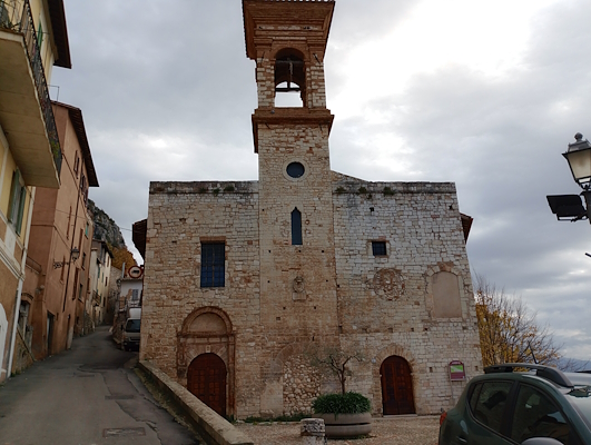 Ancient stone church with a central bell tower, located in a medieval village on a square and uphill street, under a cloudy sky.