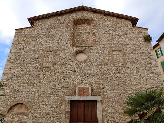 Facciata in pietra di una chiesa antica con portale ligneo e finestre murate, circondata da edifici e piante.
