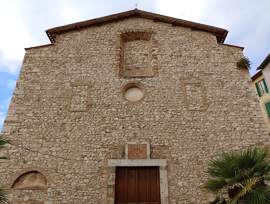 Stone façade of an ancient church with a wooden portal and walled-up windows, surrounded by buildings and plants