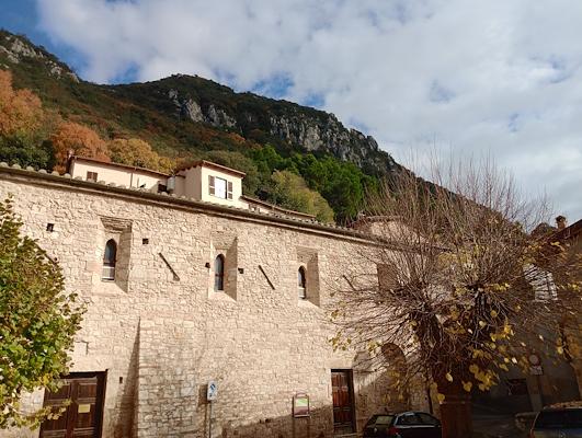 Edificio in pietra con finestre ad arco in un borgo con alta montagna alle spalle, alberi autunnali e cielo parzialmente nuvoloso.
