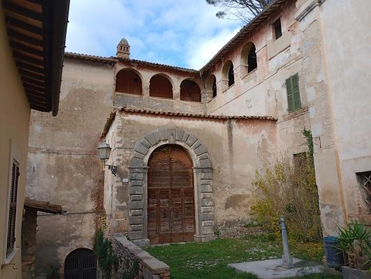 Antico edificio in pietra con grande portale ad arco e loggiato superiore, circondato da pareti intonacate e giardino erboso.