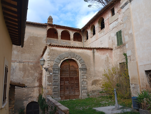Ancient stone building with a large arched portal and upper loggia, surrounded by plastered walls and a grassy garden