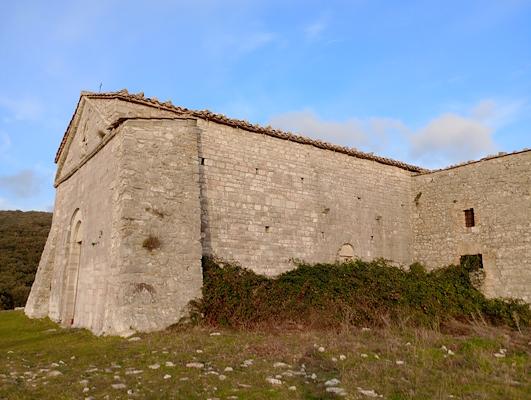 Antico edificio in pietra, circondato da vegetazione e colline, sotto un cielo azzurro con nuvole sparse.