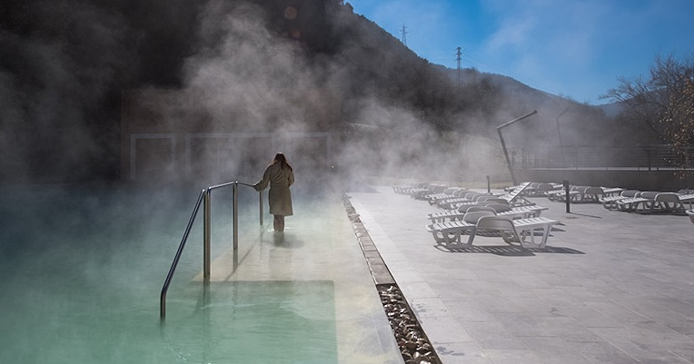Person steigt in ein dampfendes Thermalbecken im Freien, mit Bergen im Hintergrund und leeren Liegestühlen am Beckenrand
