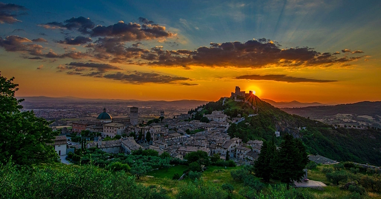 Immagine: Panoramic view of Assisi at sunset, with the sun setting behind a hill and the sky tinged with orange and blue. 