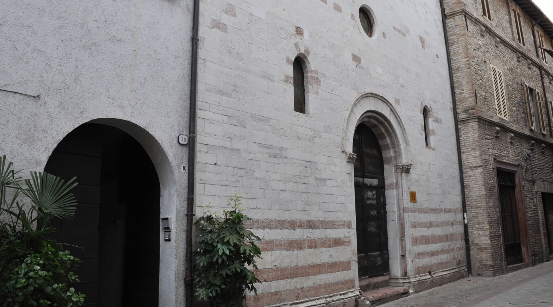 Stone façade of the Church of San Biagio in Cannara, with an arched portal and small windows, overlooking a street.